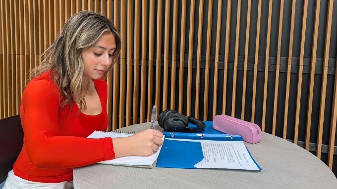 Young woman with brownish hair and a red top seated at a table studying,