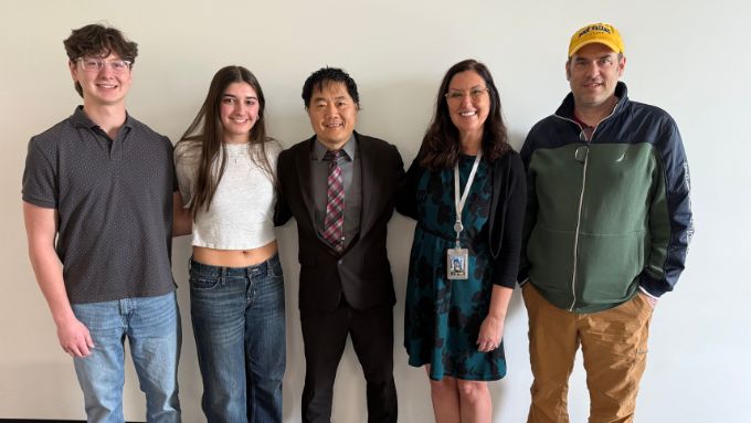 Left to right, caucasian male wearing a green shirt and jeans, caucasian female wearing a white shirt and jeans, an asian male wearing a black suit, caucasian female wearing black dress, caucasian male wearing green and black jacket and gold pants