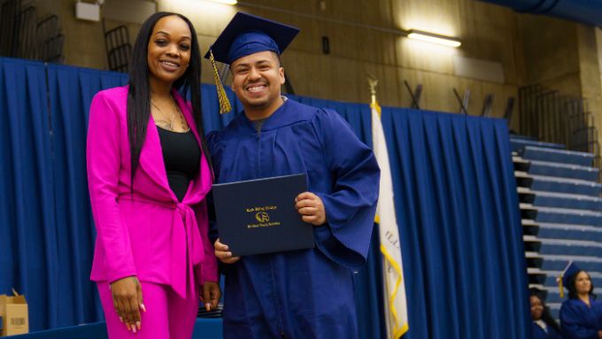 African American woman in a hot pink pants suit stands beside an Hispanic male in a blue graduation cap and gown
