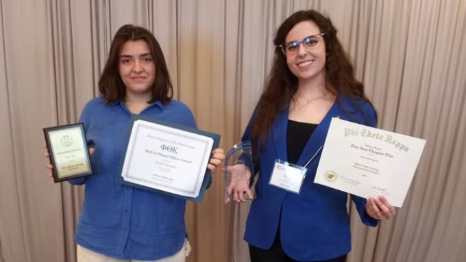 Two caucasian women wearing blue shirts. They are both holding awards received for Omicron Eta.