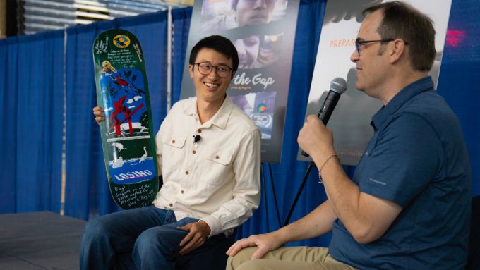Asian man (seated on left) wearing a white shirt, holding a skateboard deck talking to white man (seated on right) wearing glasses, a blue shirt and holding a microphone