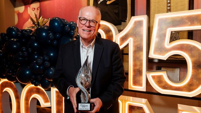 Older man holding a Citizen of the Year award with black balloons and lighted numbers in the background