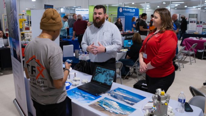 Student wesring a tan skull cap standing at an expo table with a Caucasian man and woman.