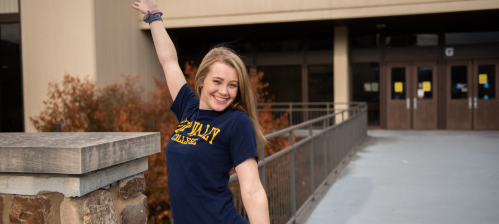 female student wearing blue RVC shirt with arms wide open on main campus bridge
