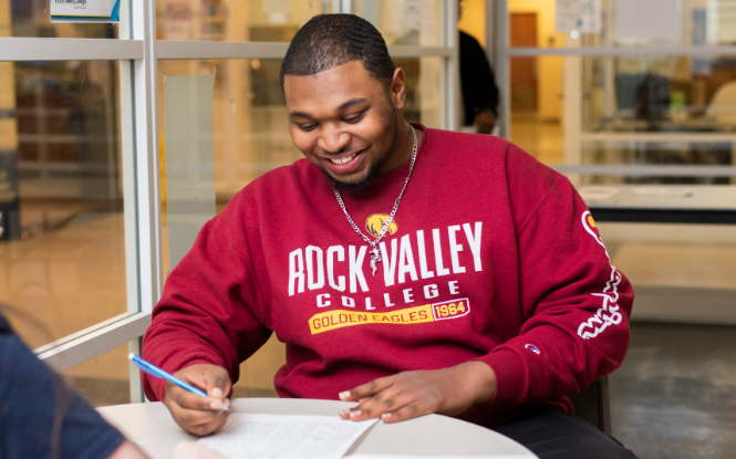 male student wearing red sweater sitting at table taking notes